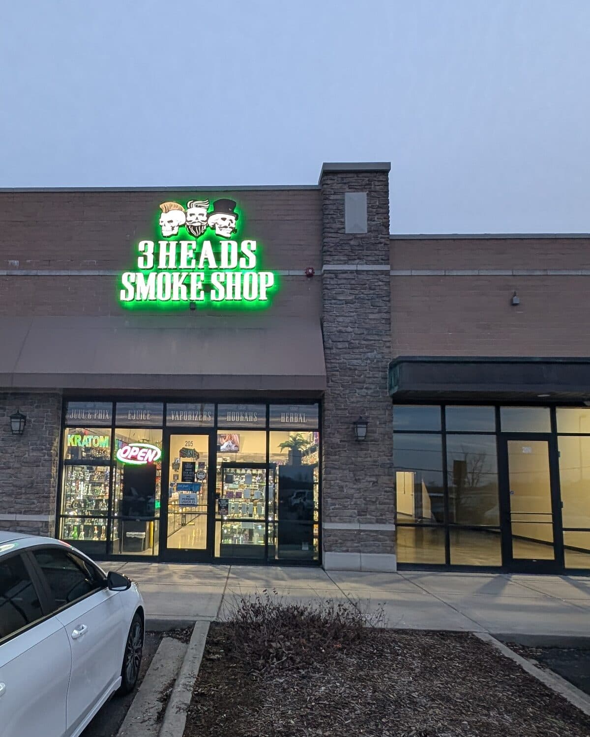 The green neon 3 Heads Smoke Shop sign with its three skulls lit up at dusk over the Elgin storefront on U.S. Route 20